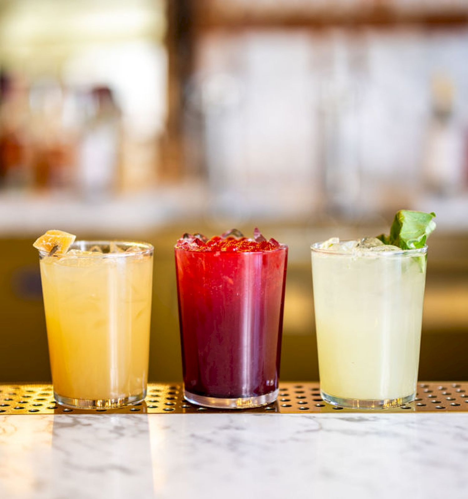 The image shows a row of three colorful cocktails lined up on a marble bar counter, each garnished differently and with varying shades of yellow, red, and green.
