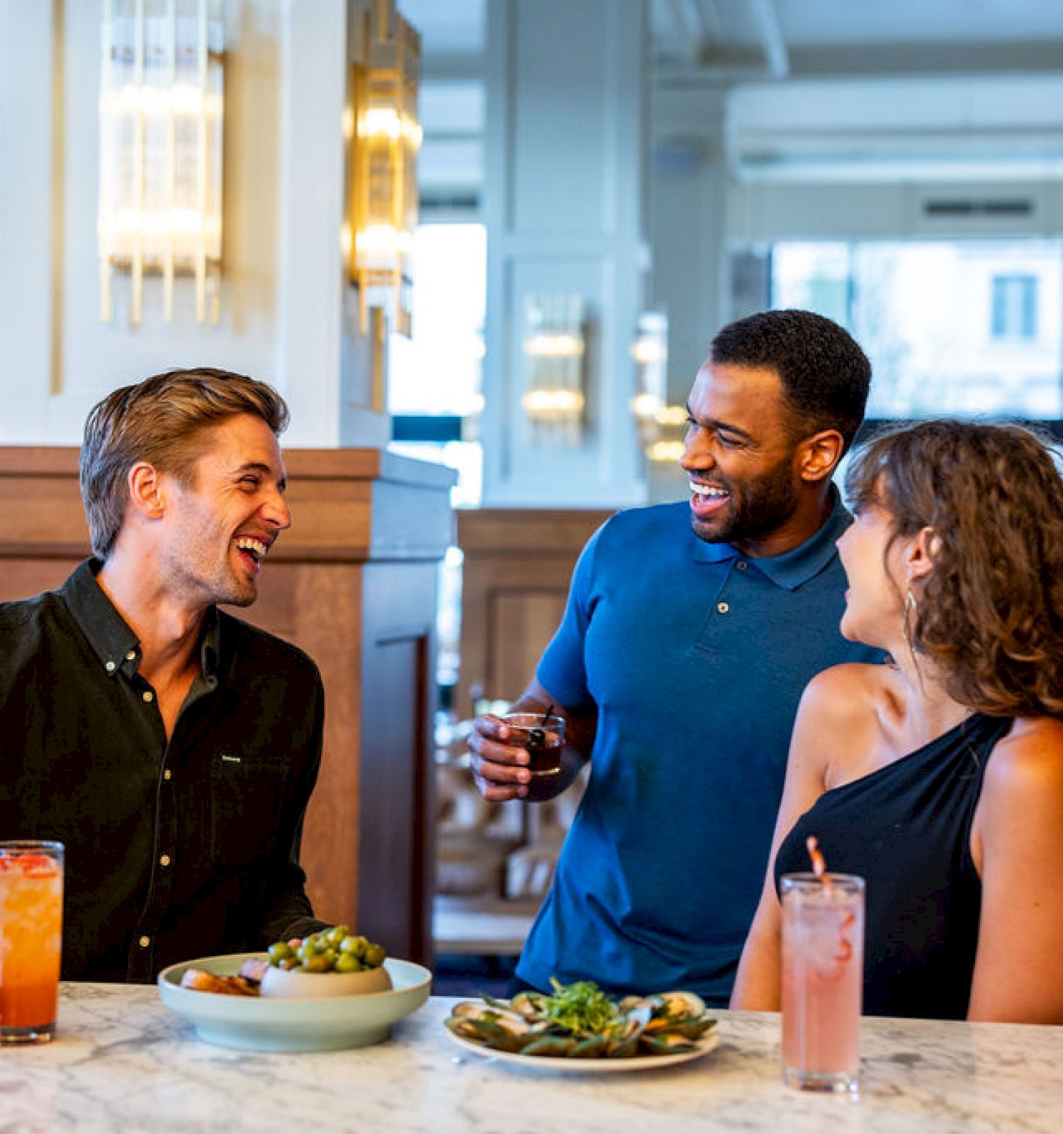 Three people are enjoying drinks and conversation at a bar or restaurant, with plates of food on the table in front of them.