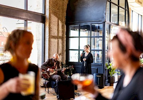 Two musicians perform indoors while two people in the foreground hold drinks and watch, with the scene having a lively atmosphere.
