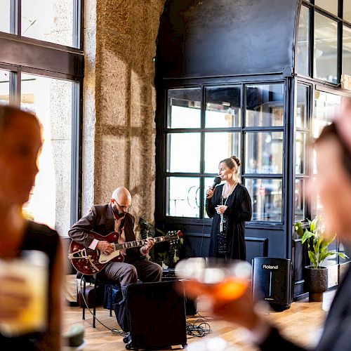 Two musicians perform indoors while two people in the foreground hold drinks and watch, with the scene having a lively atmosphere.
