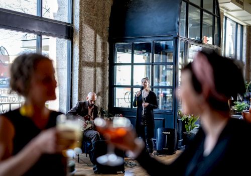 People are gathered in a restaurant or bar. Musicians perform in the background as two women clink glasses in the foreground, enjoying their drinks.