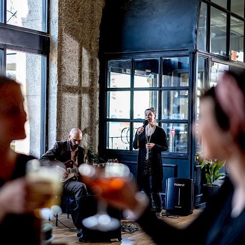 People are gathered in a restaurant or bar. Musicians perform in the background as two women clink glasses in the foreground, enjoying their drinks.