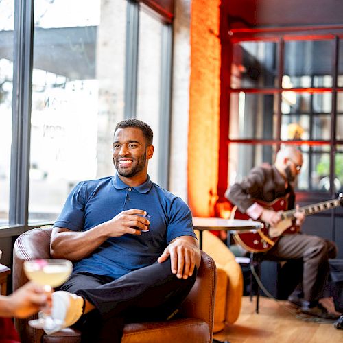 People are relaxing in a cozy lounge, enjoying drinks and live music performed by musicians playing the guitar and saxophone.