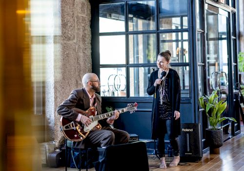 A man is playing an electric guitar, and a woman is singing into a microphone in a well-lit indoor space with large windows and greenery.