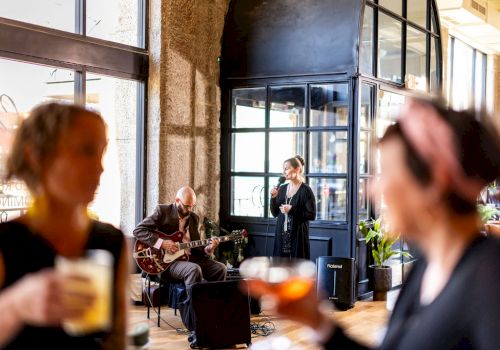 A small musical performance is happening with a singer and a guitarist, while two blurred individuals in the foreground hold drinks.