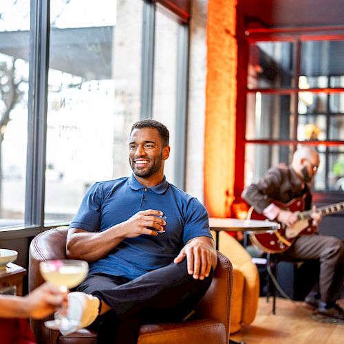 People are relaxing in a stylish lounge; a man enjoys a drink while musicians play in the background, with a woman seated nearby.