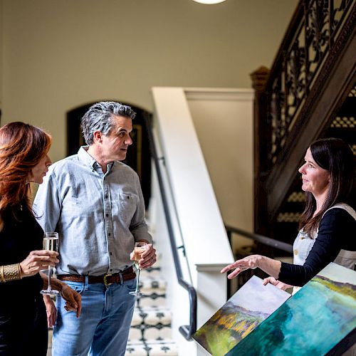 Four people are gathered by a staircase, engaging in conversation, with two holding drinks and one pointing towards an art piece on an easel.