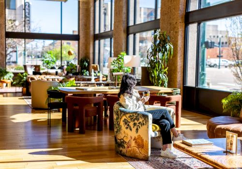 A woman is sitting in a cozy, brightly lit room with large windows, surrounded by plants and furniture, enjoying a beverage and looking outside.