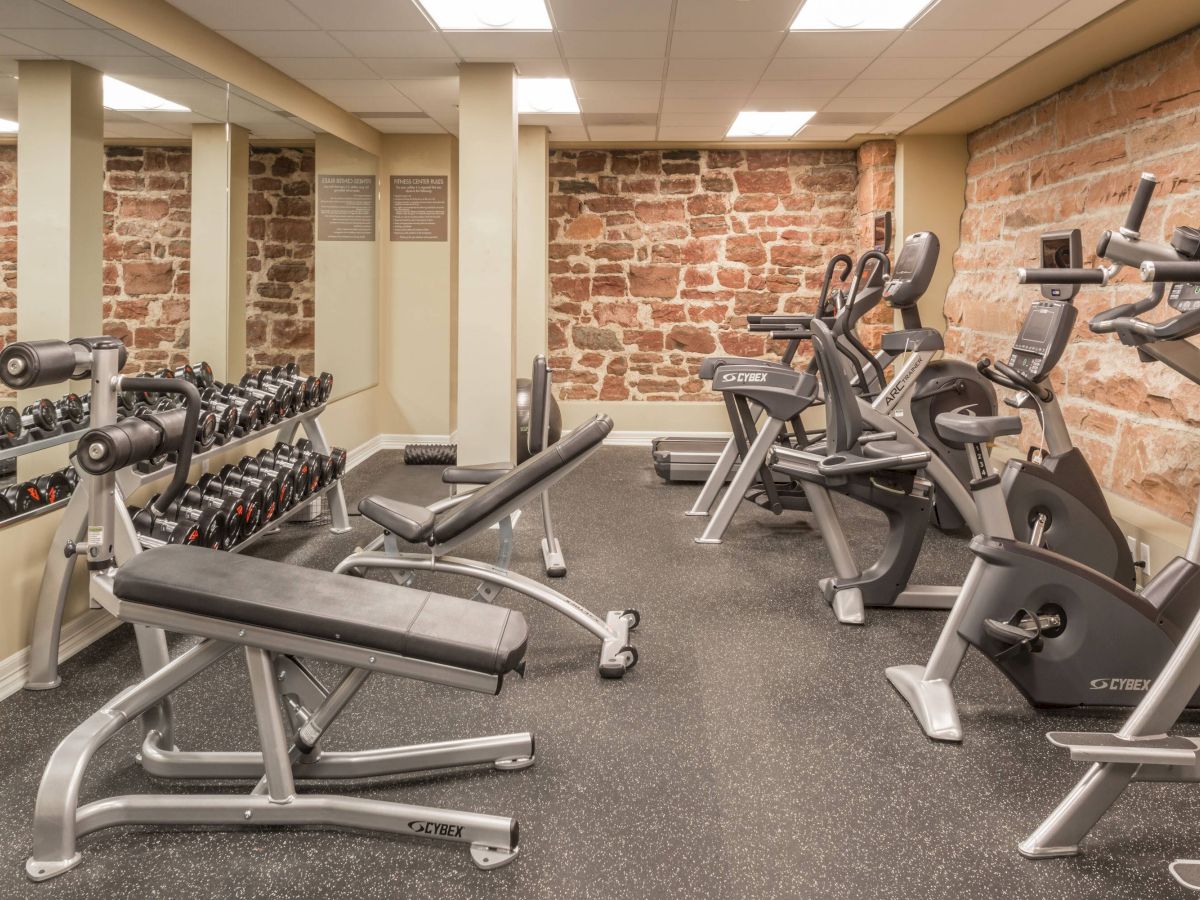 A well-equipped gym with weights, benches, and cardio machines against a backdrop of stone walls and wall-length mirrors.