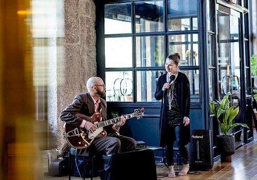 A guitarist and a singer are performing indoors; the guitarist is seated and the singer is holding a microphone, with a modern interior backdrop.