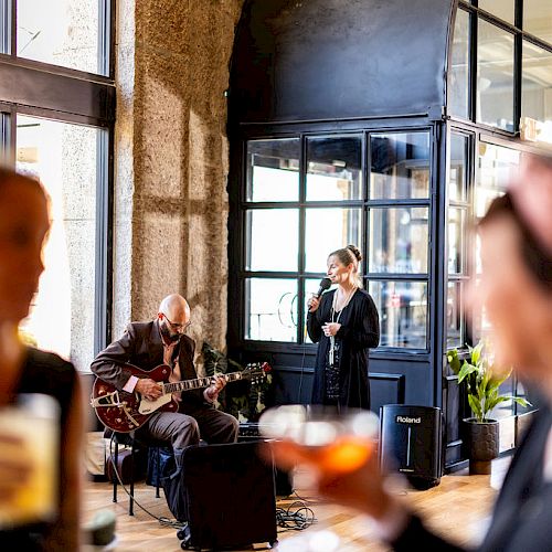 A musician plays guitar while a singer performs at a venue with blurred people in the foreground holding drinks.