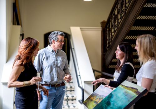 Four people are gathered indoors near a staircase, seemingly engaged in conversation, with one displaying a piece of artwork.