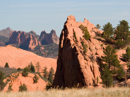 The image shows a scenic view of red rock formations, mountains in the background, and scattered green trees under a clear blue sky.