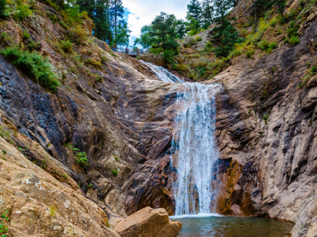 A beautiful waterfall cascades down rugged cliffs surrounded by lush greenery, with a pool of water at the base and trees lining the rocky terrain.