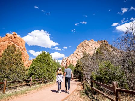 A couple walks on a scenic path surrounded by red rock formations and greenery under a bright blue sky with scattered clouds, capturing a peaceful moment.