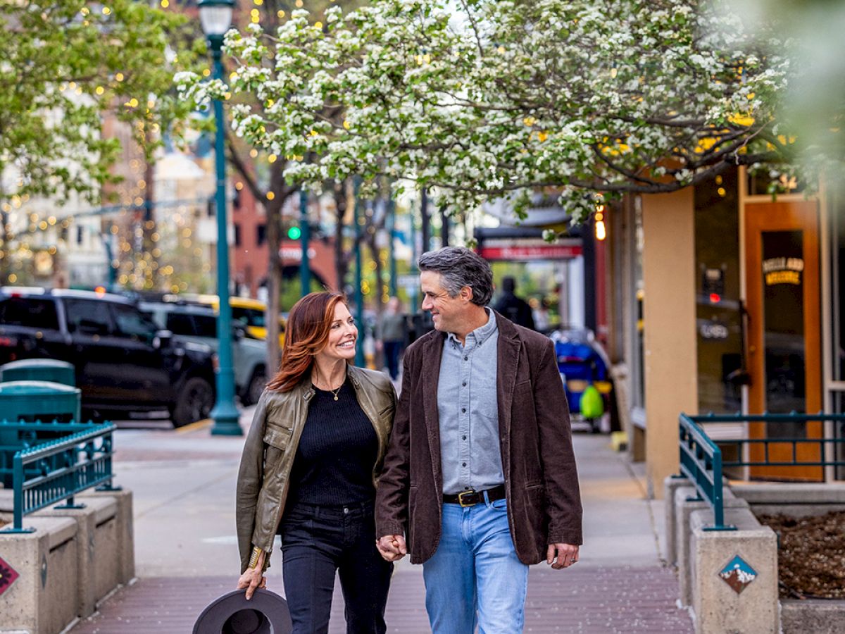 A couple is walking hand in hand on a tree-lined street, smiling at each other. The man is holding a hat, and there are shops and greenery around.