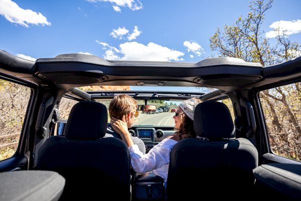 A couple enjoys a drive in a convertible under a clear blue sky, with trees and a road stretching ahead.