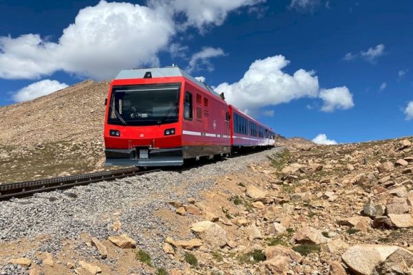 A red and white train travels on tracks through a rocky, mountainous landscape under a partly cloudy sky.