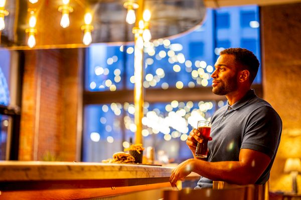 A person is standing at a bar holding a drink with a festive, illuminated background, featuring bokeh lights.