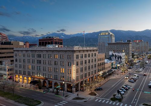 An aerial view of a city at dusk, showcasing buildings, streets with vehicles, pedestrians, and distant mountains under a twilight sky.