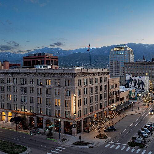 An aerial view of a city at dusk, showcasing buildings, streets with vehicles, pedestrians, and distant mountains under a twilight sky.