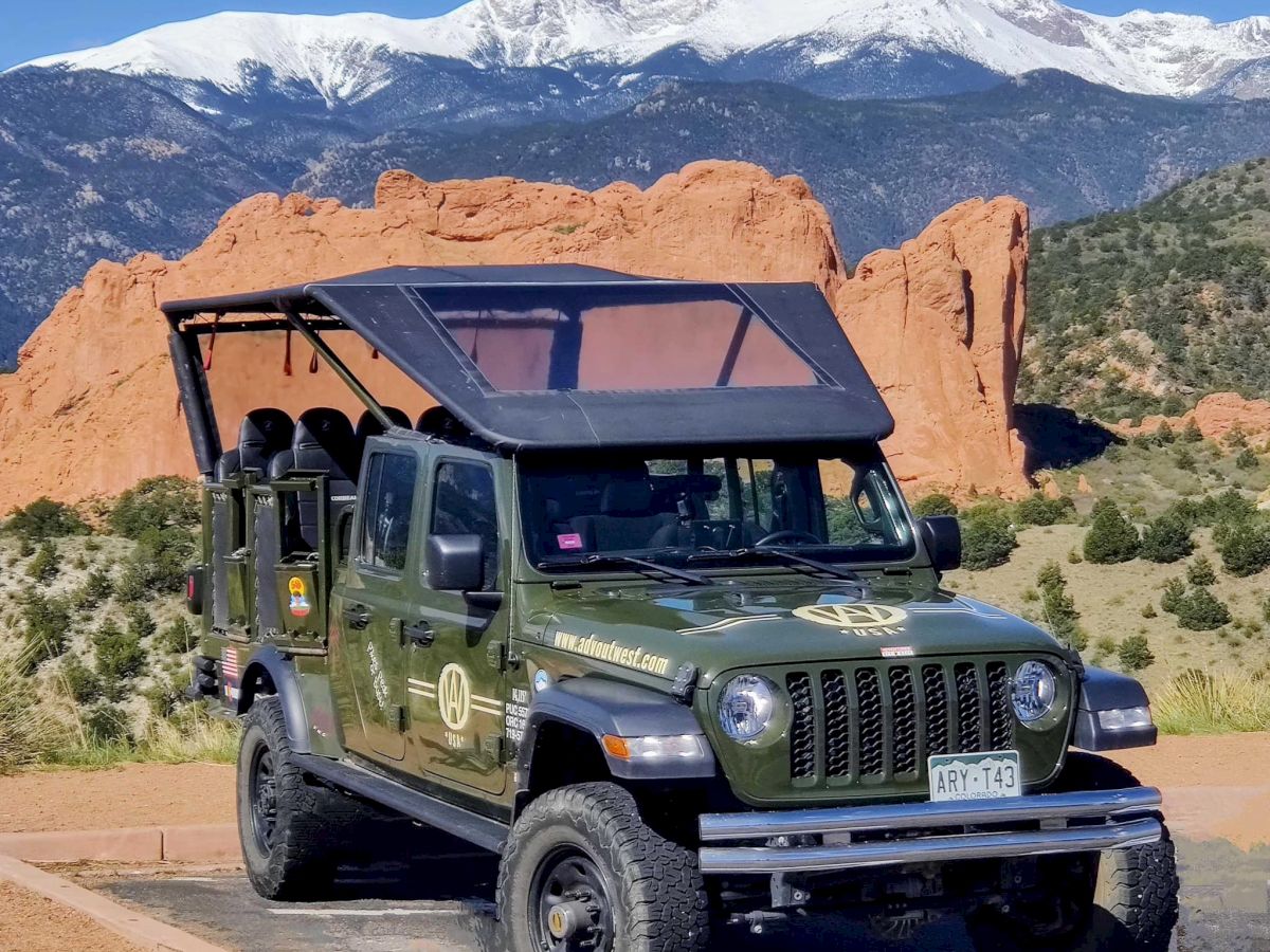 An off-road vehicle with a partially open roof is parked in front of a scenic landscape featuring mountains and red rock formations, under a clear sky.