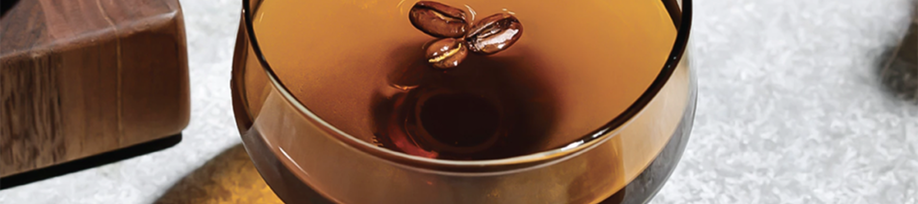 A cocktail glass filled with a dark beverage, garnished with two coffee beans on top, placed on a light-colored surface.