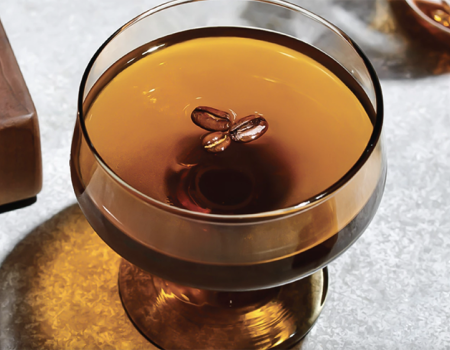 A cocktail glass filled with a dark beverage, garnished with two coffee beans on top, placed on a light-colored surface.