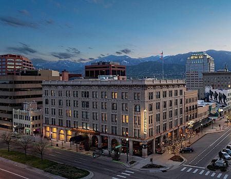 This image shows a city street at dusk with several buildings, cars on the road, and mountains in the background. The scene includes streetlights and a calm atmosphere.