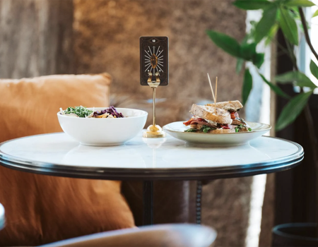 A table in a cozy setting with a bowl of salad, a sandwich, and a small card in a holder, next to cushioned seating and plants.