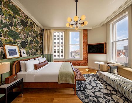 The image shows a stylish hotel room with a bed, a unique wallpaper, large windows, a sofa, a TV, and modern decor, creating a cozy atmosphere.