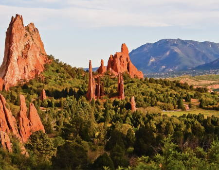 The image shows a landscape with towering red rock formations surrounded by lush greenery, with a mountain range in the background and a bright sky.