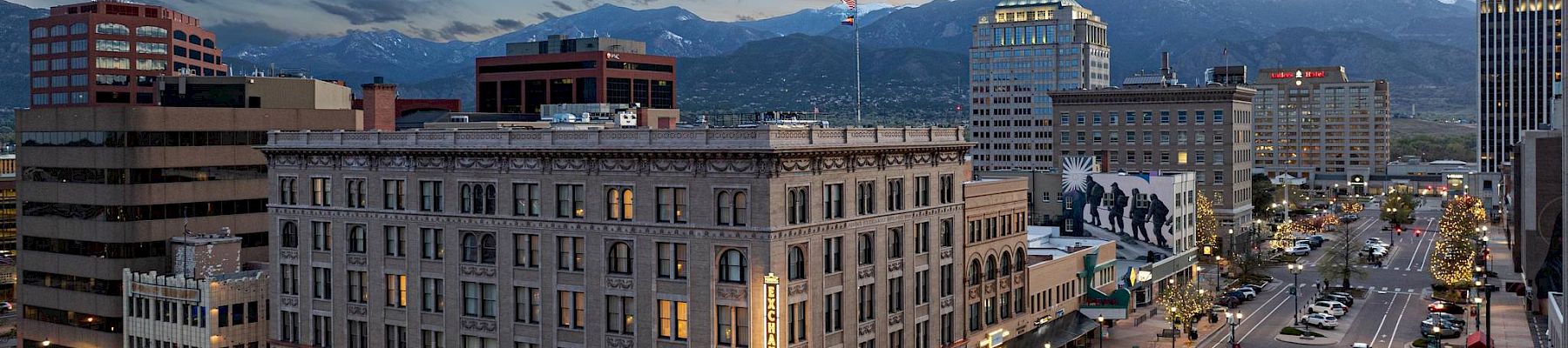 A cityscape at dusk featuring buildings, streets with cars, and a backdrop of mountains under a twilight sky.