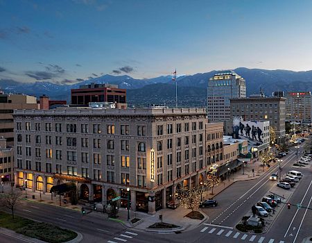 A cityscape at dusk featuring buildings, streets with cars, and a backdrop of mountains under a twilight sky.