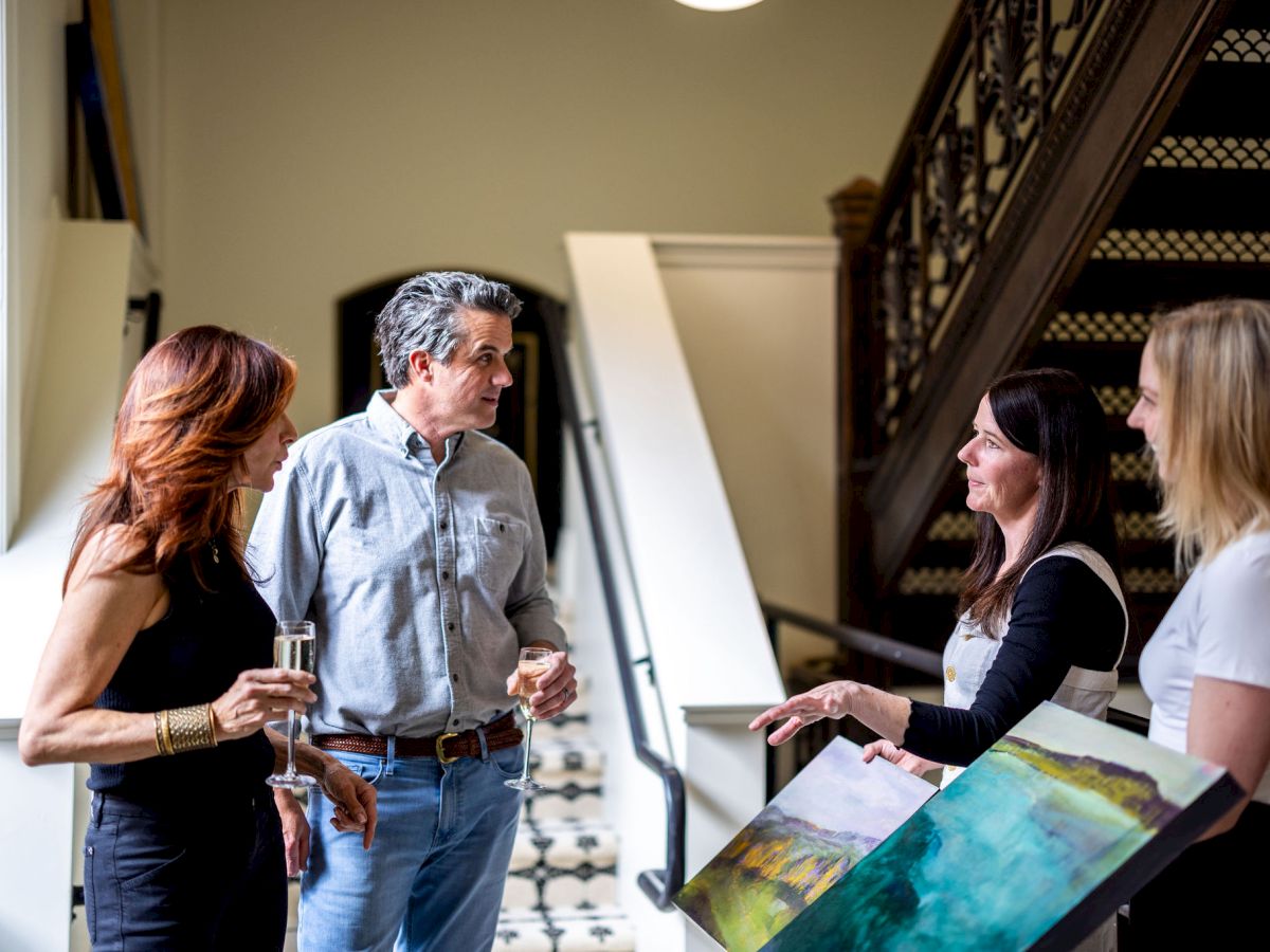 A group of people are conversing near a staircase, with one person holding a painting. They appear to be in a formal or social setting.