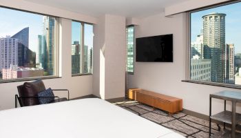 The image shows a modern room with large windows offering a city view, a bed, a wall-mounted TV, a chair, and a small table.