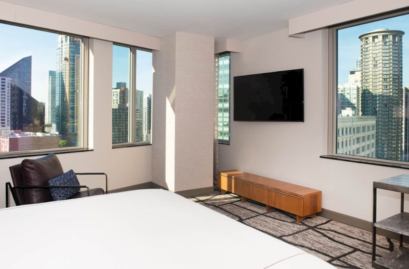 The image shows a modern room with large windows offering a city view, a bed, a wall-mounted TV, a chair, and a small table.