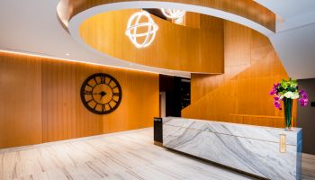 A modern reception area with wooden panels, a marble desk, a large wall clock, a spiral staircase, and floral arrangements on the desk ends the sentence.