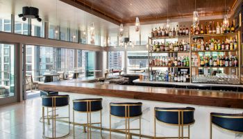 A modern bar with a wooden counter, high-back stools, and a well-stocked shelf of various liquor bottles. Large windows offer a city view.