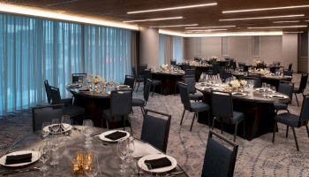 The image shows an elegant, dimly-lit banquet hall set with round tables, black chairs, tableware, and floral centerpieces, ready for an event or dinner.