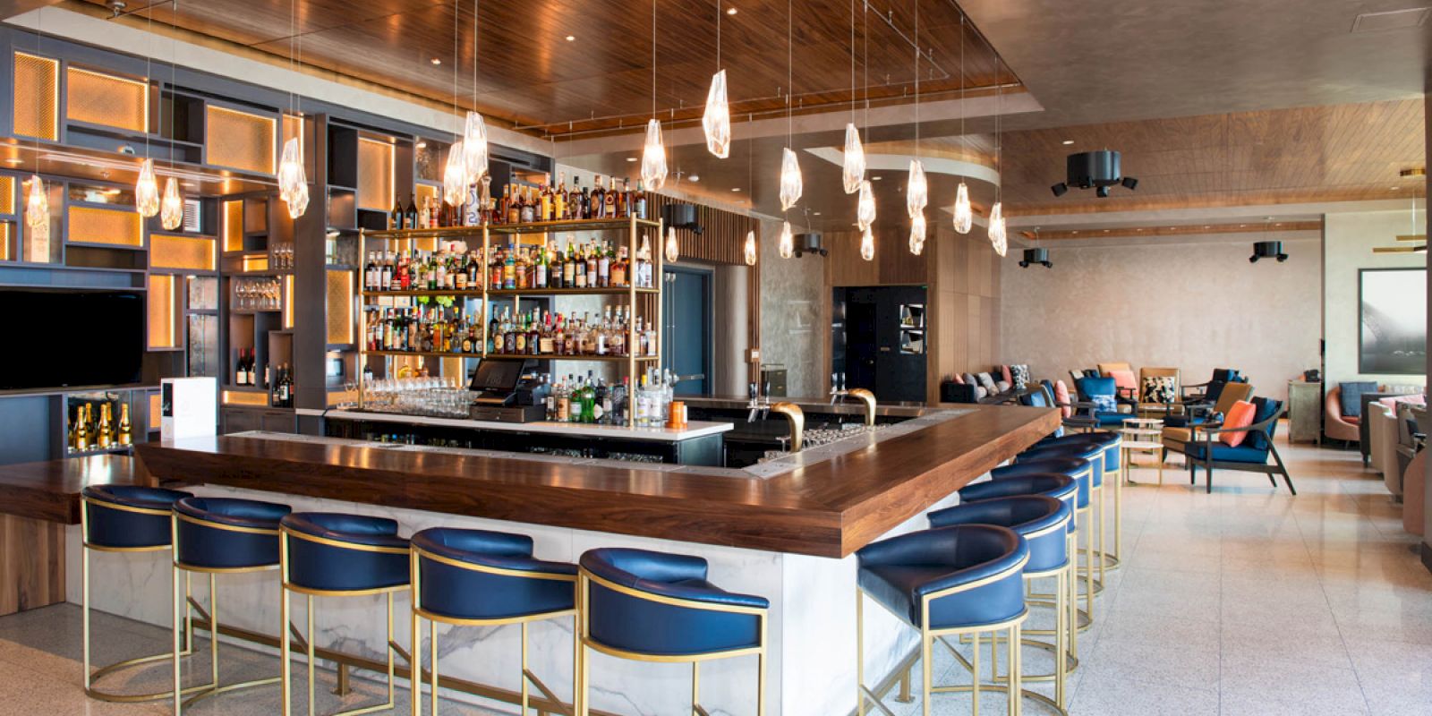 A modern bar area with a well-stocked liquor shelf, stylish lighting fixtures, and blue stools. Adjacent lounge with seating.
