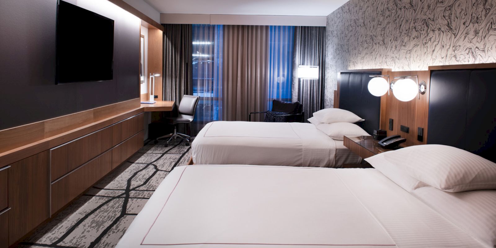 A modern hotel room with two beds, wall-mounted TV, desk, chair, and lamps. The room features minimalist decor with abstract wallpaper and furnishings.