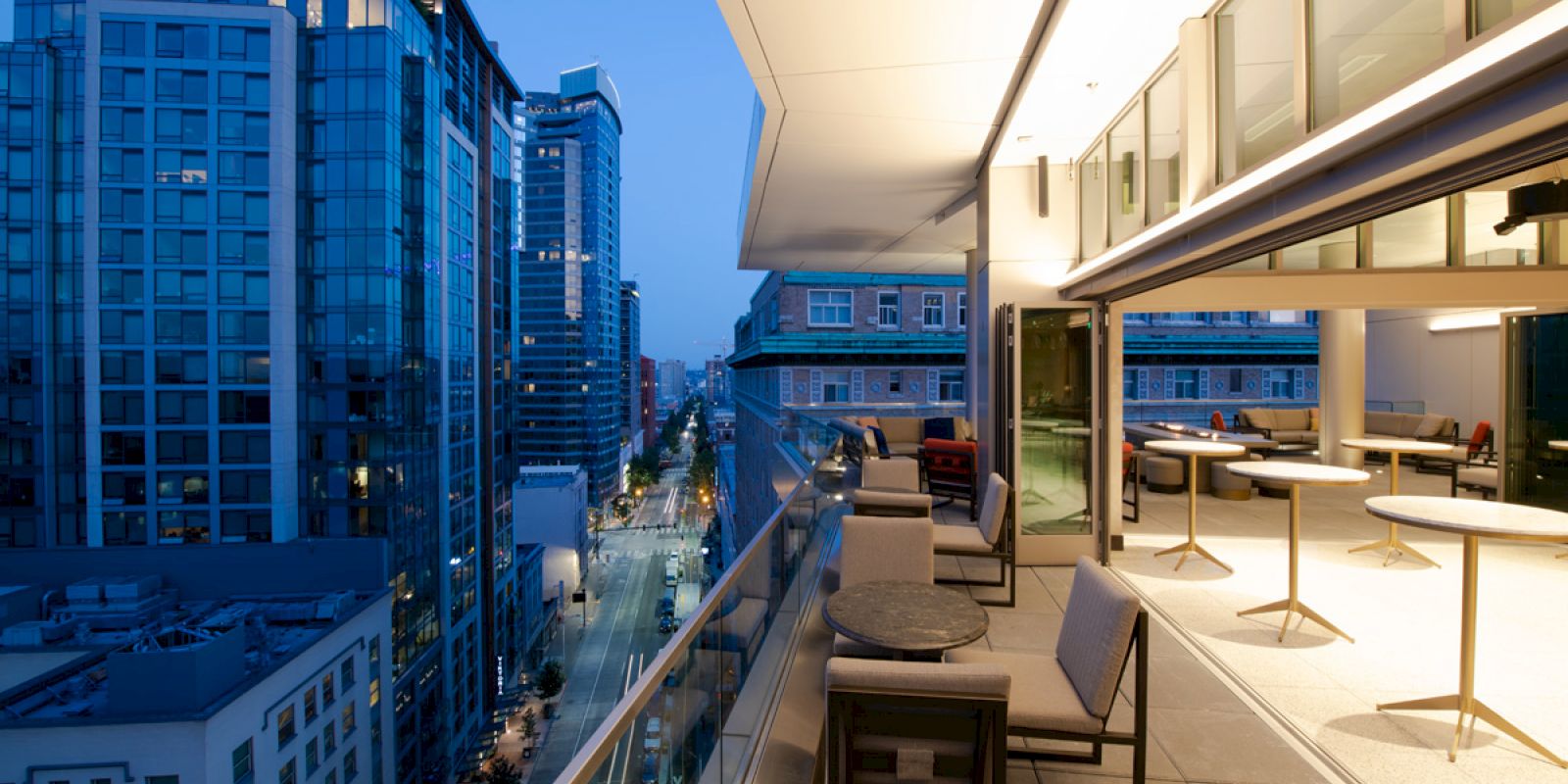 A modern balcony with chairs and tables overlooks a cityscape with high-rise buildings at dusk, featuring floor-to-ceiling windows and ample lighting.