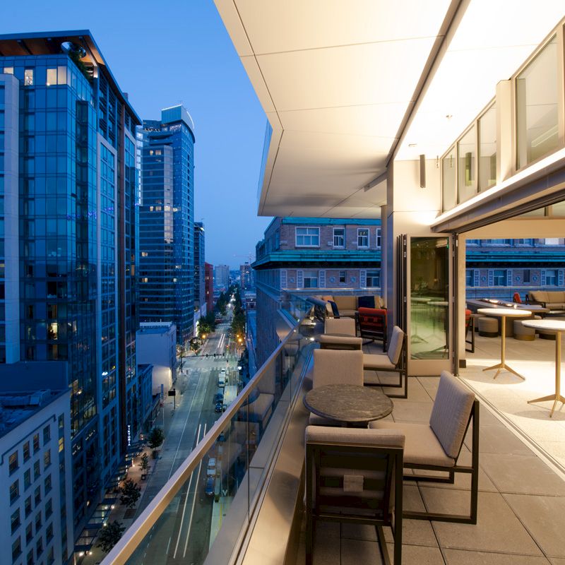 A modern cityscape view from a high-rise balcony, featuring tables and chairs, with buildings and a street visible at dusk, under a blue sky.