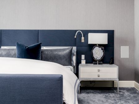 A modern bedroom features a navy blue upholstered headboard, white bedding, a bedside table with a lamp, and gray carpet flooring.