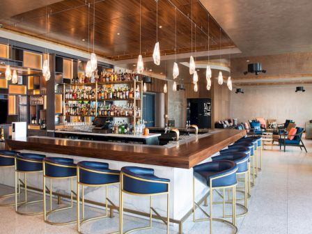 The image shows a modern bar with high chairs neatly arranged around a wooden counter, shelves stocked with bottles, and pendant lights hanging.