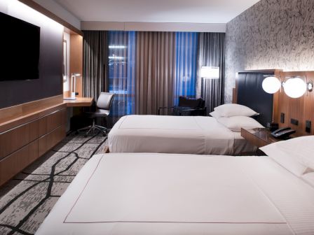 A modern hotel room with two beds, a TV, a work desk, a chair, lamps, and curtains. The room features contemporary decor and a stylish design.