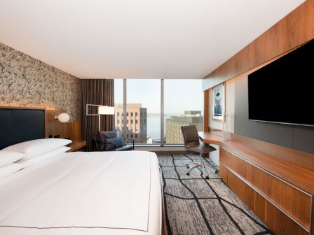 A modern hotel room with a large bed, wall-mounted TV, desk with chair, and a window offering a cityscape view, including buildings and water.