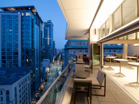 A modern cityscape with tall buildings viewed from a stylish, furnished balcony at twilight, featuring tables, chairs, and interior lighting.