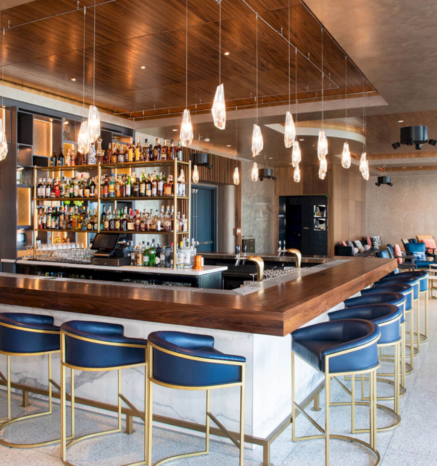 A stylish bar with a wooden counter, blue upholstered chairs, overhead lighting, and a fully-stocked bar area, set in a modern lounge environment.